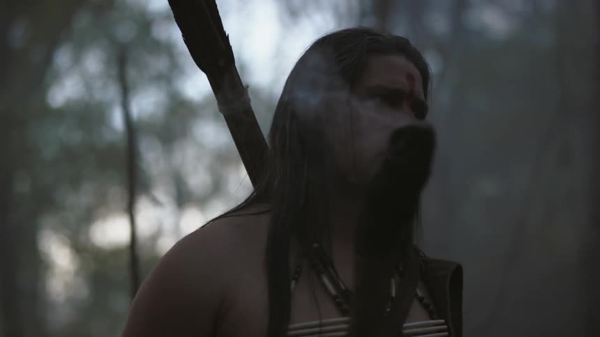 A native american carrying weapons and a smoking torch creeps through a forest