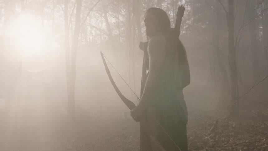 Man stands holding bow as the sun shines through trees