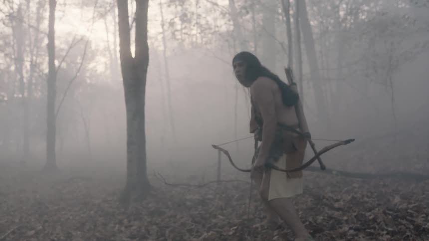 A native american carrying a bow and arrow quietly walks through a misty forest
