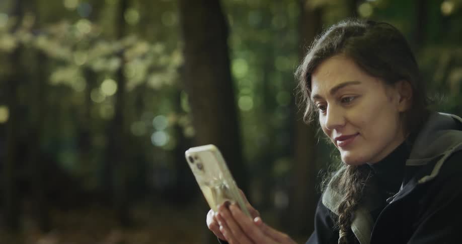 A woman holds her smart phone and smiles as she is located within a forest