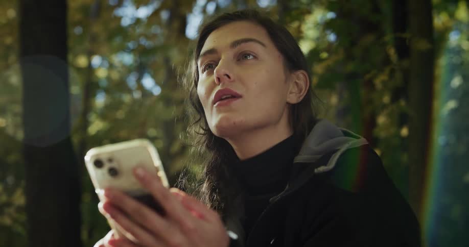 A woman is in the woods and she takes off her cycling helmet and holds her cell phone in her hand