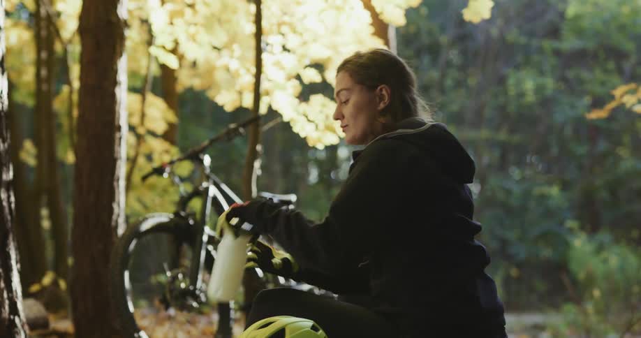 A young woman sits in a forest by her bike and takes her helmet off and takes a drink from her water bottle