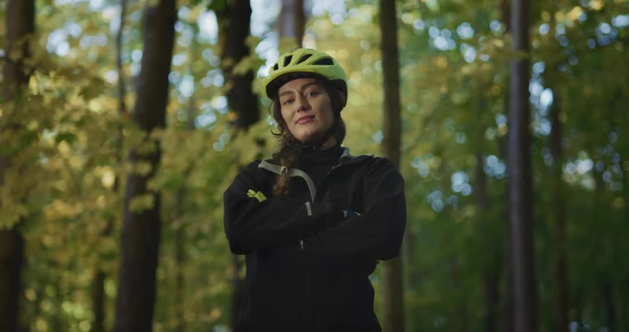 A woman with a bike helmet on stands in a forest and smiles