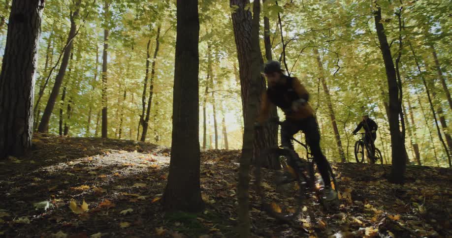 Two cyclist ride down fallen leaf strewn hill onto pavement in woods