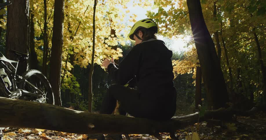 A woman wearing a cycling helmet is sitting on a log in the woods near her bike and holding her phone as the sun shines brightly through the trees