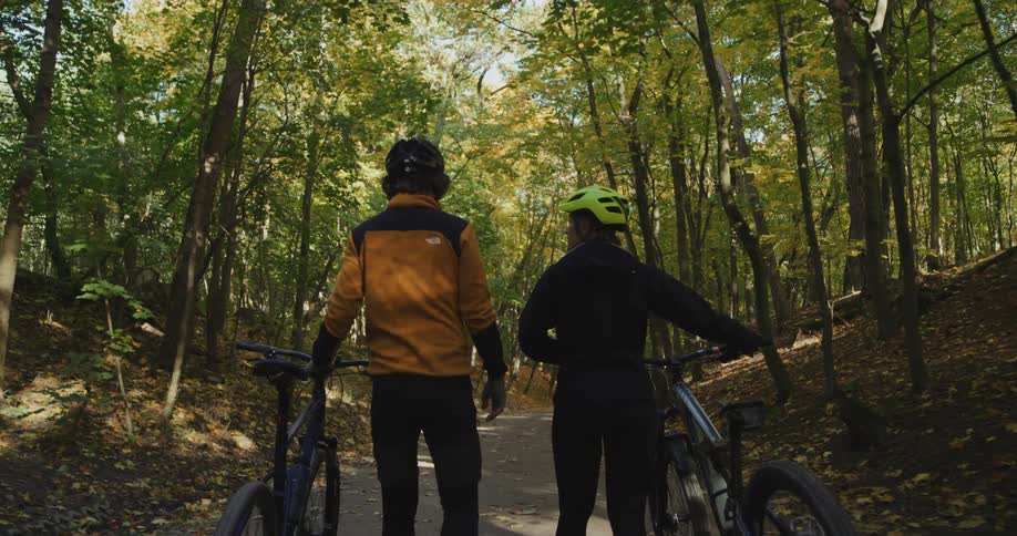 Two bicycle riders walk along a forest trail while rolling their bicycles next to them