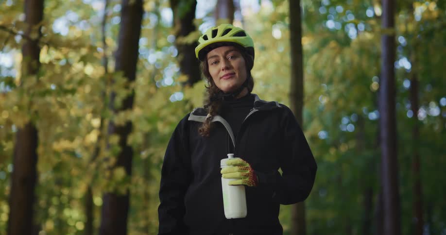 A woman wearing a cycling helmet is standing in the woods holding a water bottle