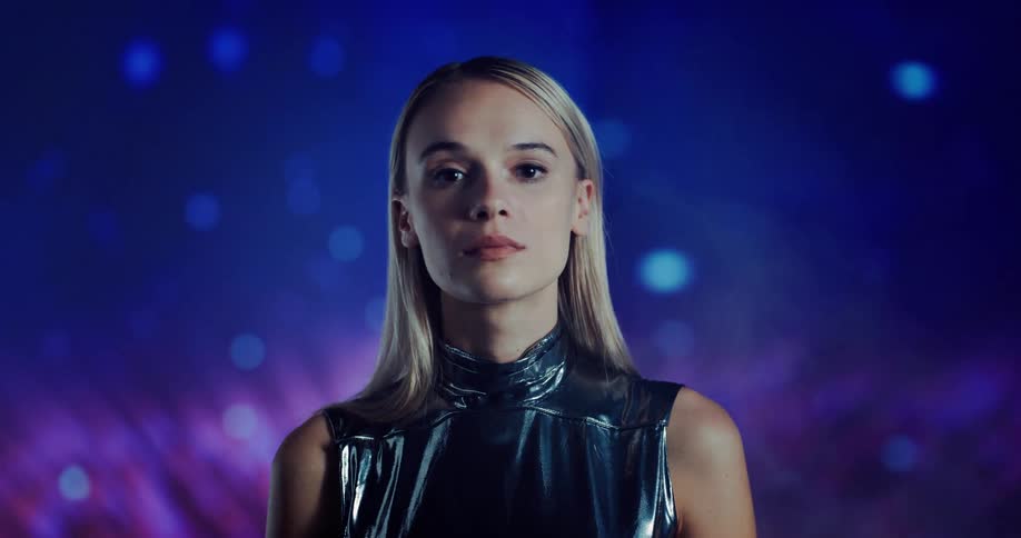 Young blonde woman wearing silver reflecting clothes stands inside a room with lines projected on a wall