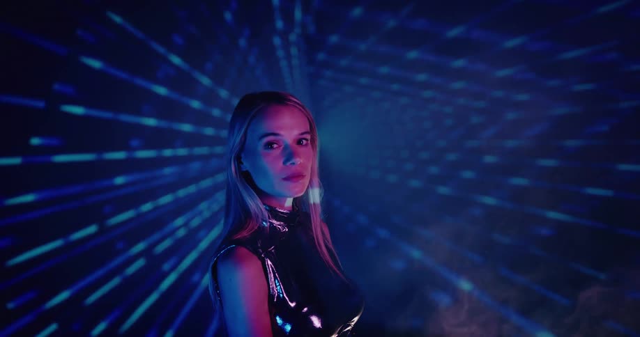 Blonde woman wearing metallic sleeveless outfit stands and stares as blue light beams create zooming effect