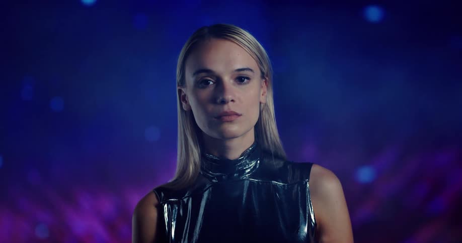 A woman with long blond hair is standing in an environment with moving purple and pink lights behind her and she is looking down and then she looks straight ahead