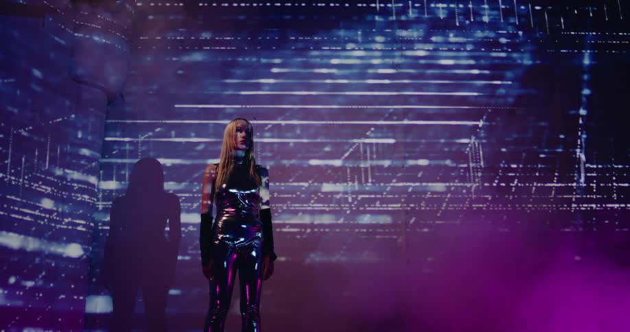 Young blonde woman wearing silver reflecting clothes stands inside a room with lines projected on a wall