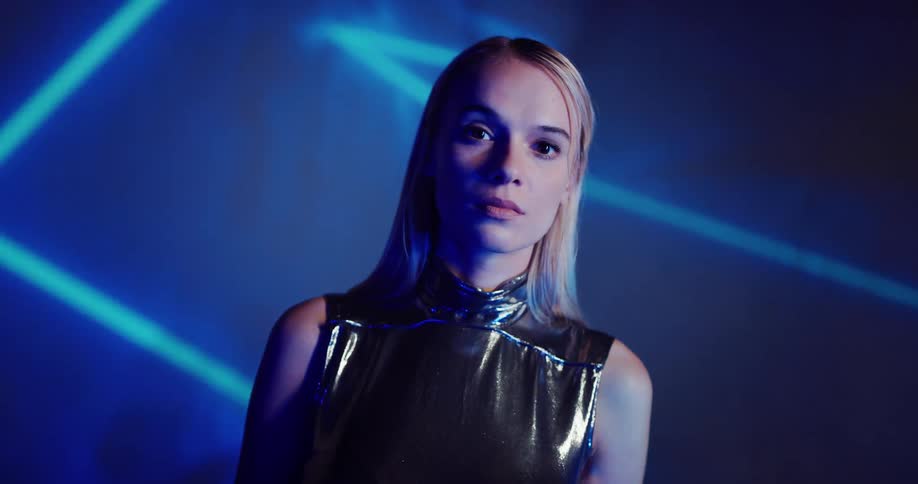Blonde woman stands and stares as colored lights create shapes behind her