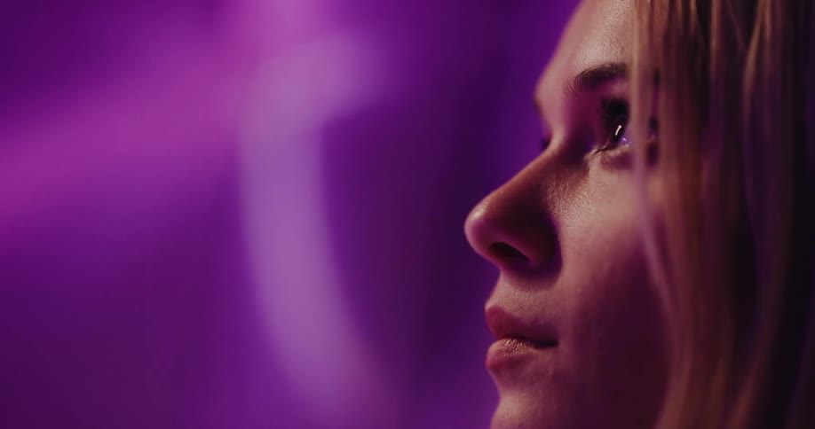 Close up of the face of a young beautiful woman standing on a room under projected blue and purple lights