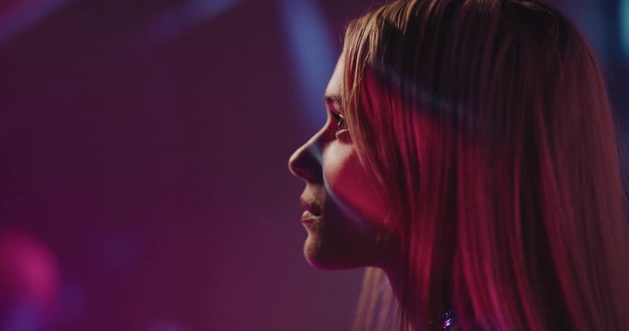 A woman with long blond hair is standing in a room with moving pink colored lights as they shine on her face