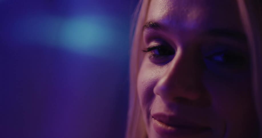 Close up of the face of an attractive blonde woman standing on a room under colorful lights looking at the camera
