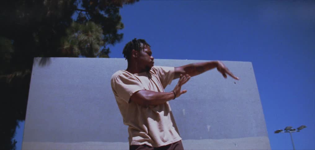 A man is standing outside in front of a white board dancing and moving his arms and hands under a clear blue sky