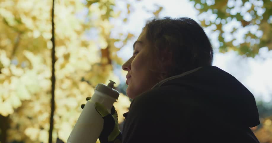 Female cyclist takes off bicycle helmet and drinks from reusable water bottle under trees on a sunny day
