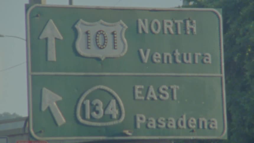 California road sign stands near trees as cars pass on overpass in the background