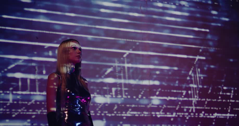 Young blonde woman wearing silver reflecting clothes stands inside a room with lines projected on a wall