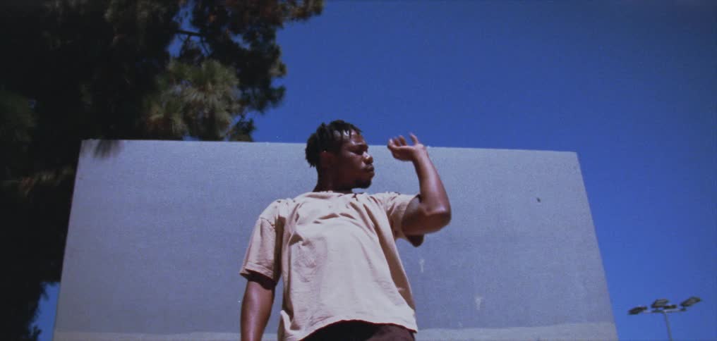 A man is standing outside in front of a white board doing hand and arm dance movements under a clear blue sky