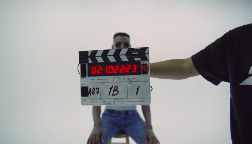 Assistant claps a board in front of a model sitting on a bench inside a studio