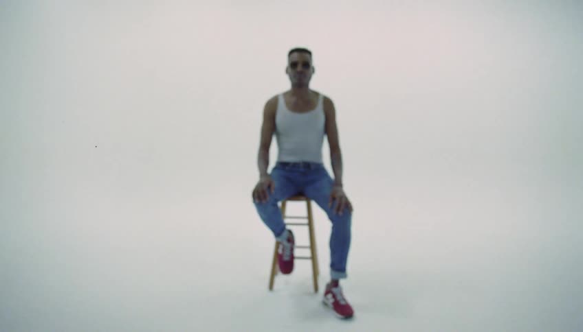 A man holds out a clapperboard with moving digital numbers counting down as a man wearing sunglasses is sitting on a stool