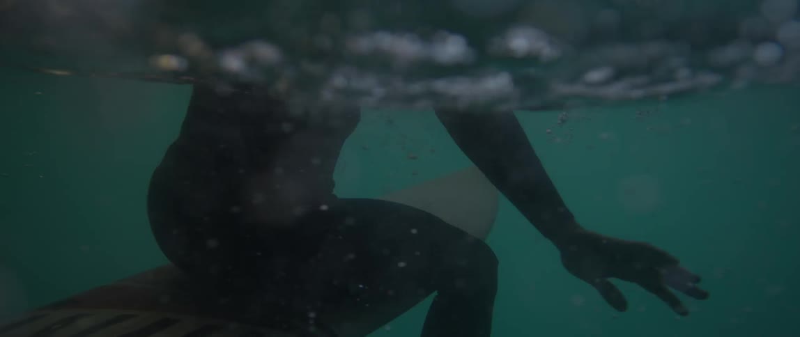 A surfer sits on his surfboard in the water as seen from below the surface
