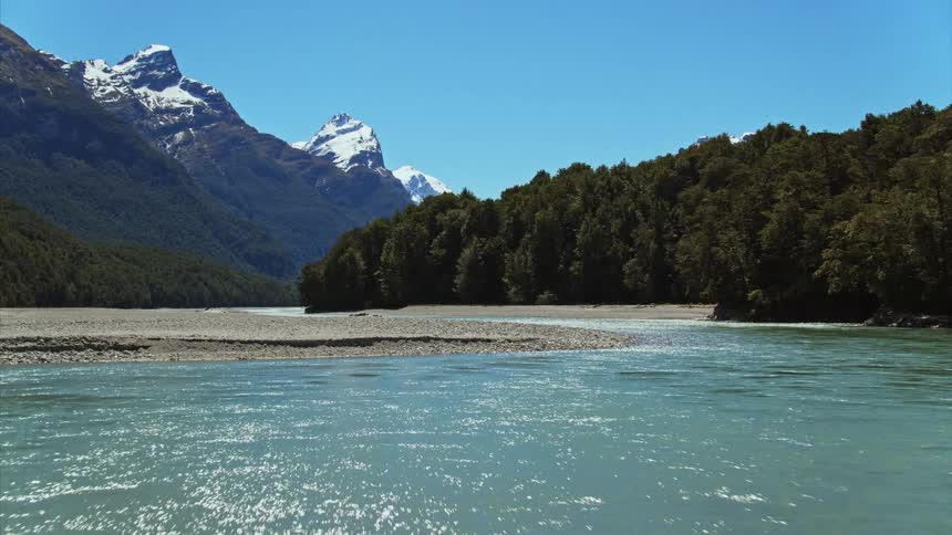 Low level flying across clear blue alpine river in front of a mountain range
