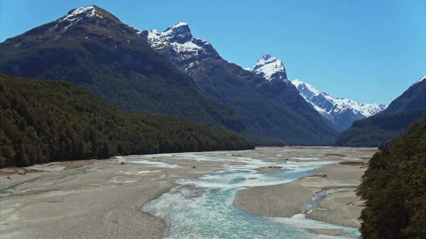 An aerial track past historic trees to reveal blue braided river and dramatic mountain range