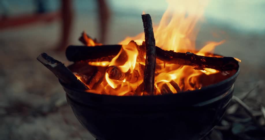 A woman on a beach stands behind a fire burning in a pit