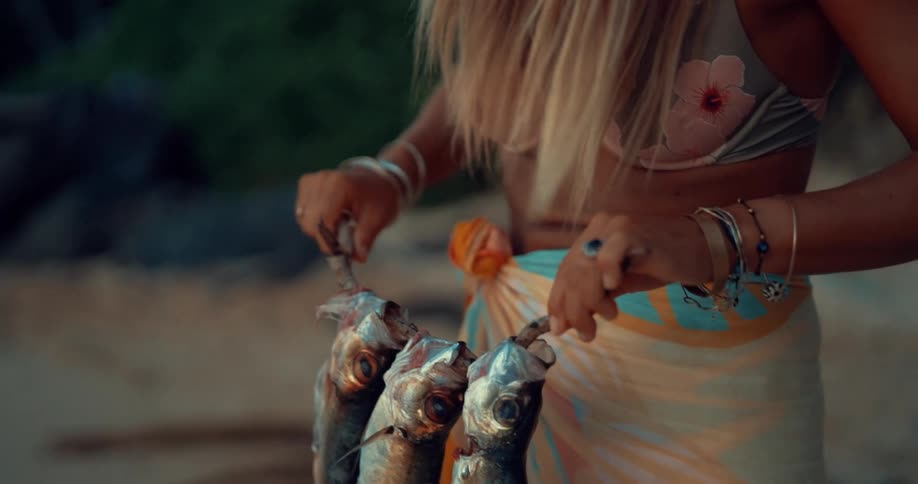 Woman wearing bikini spits three fish on a branch to cook