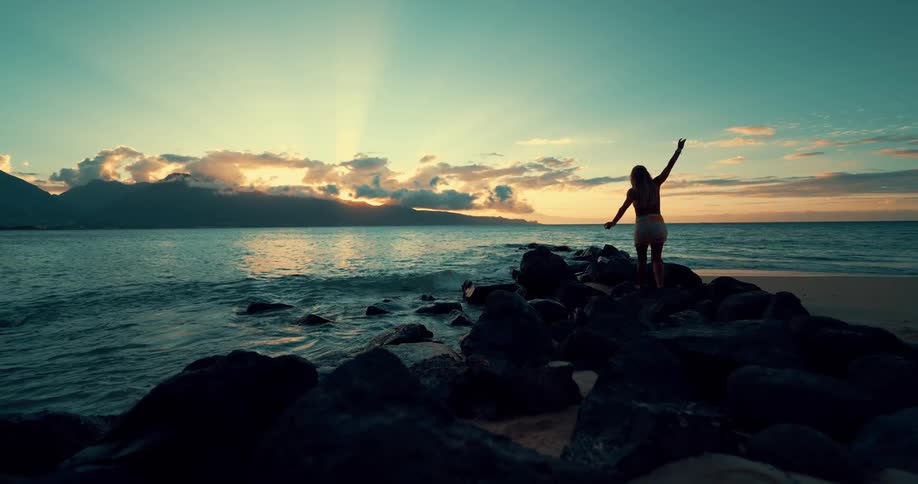 A woman walks over a rock jetty toward the ocean at sunset
