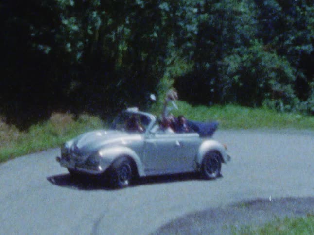 People are riding in a convertible volkswagen as they go around a curve on the road