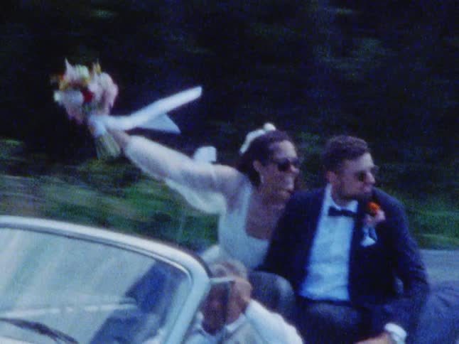 A bride and groom sit in the back of a convertible as someone drives and they kiss