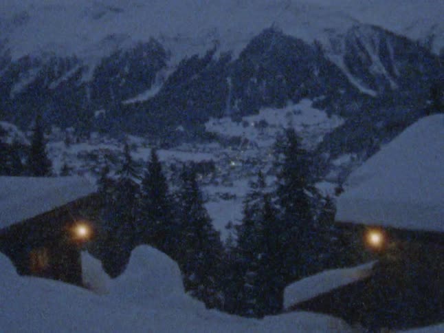 Light of town buildings shine dimly as snow falls onto buildings and mountains