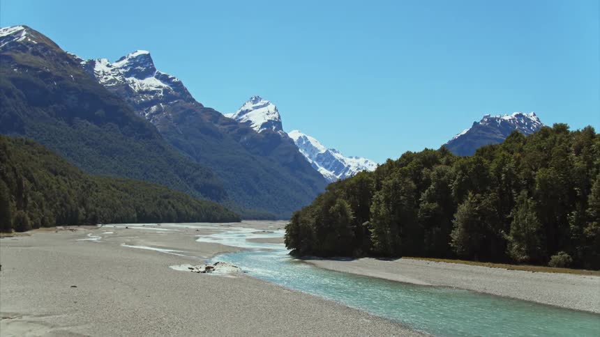 Track back along light blue water flowing past conifer forest with snowy mountains in the background