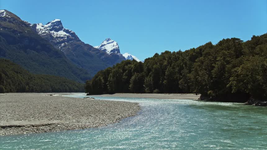 Light blue water flows along channel past sand and pebble shore and conifer forest toward snowy mountains