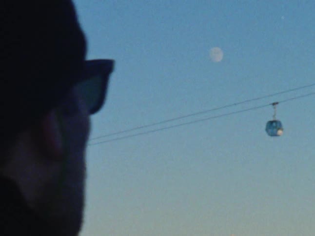 Man watches two overhead cable cars pass each other near mountains under the moon during the day