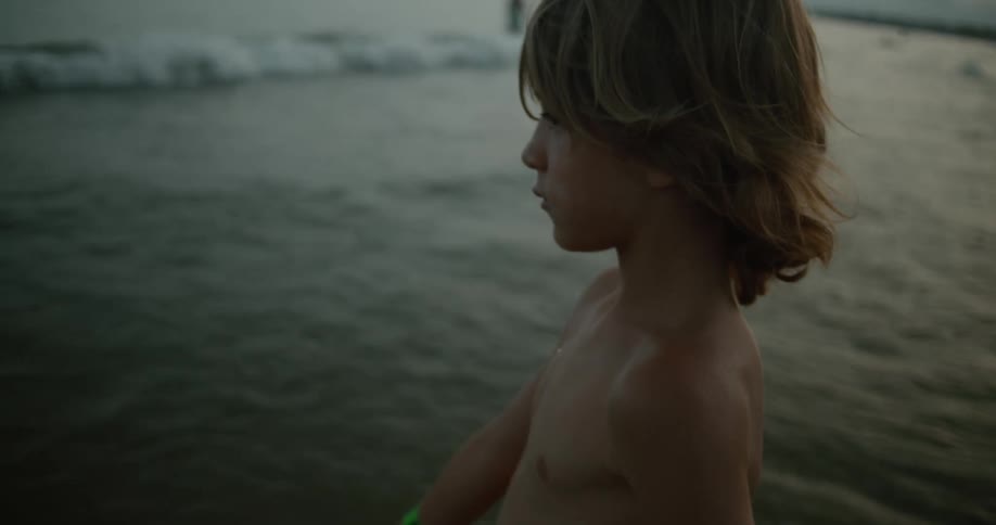 A shirtless boy walking into ocean waves on a beach at sunset
