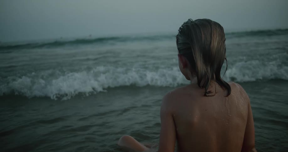 A boy is sitting in the water at the beach as waves roll into shore and splash on his chest