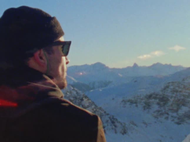 A man is standing on a mountain ledge looking at the valley below and snow covered mountains in the distance