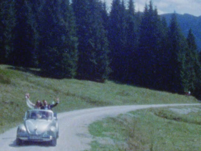 A bride and groom standing and waving in a convertible while driving on a dirt road