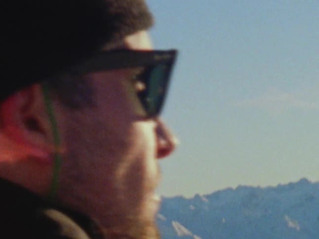A man in a winter cap and sunglasses looking out at snowy mountains