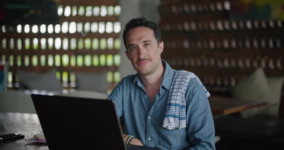 A man is sitting at a table behind a laptop computer with a towel over his shoulder