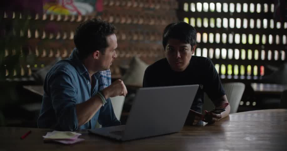 Two men sit at a table in an office and look at a computer and talk