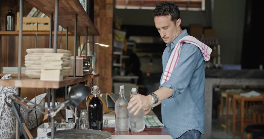 A male restaurant employee slinging a towel over his shoulder and picking up glass bottles of water