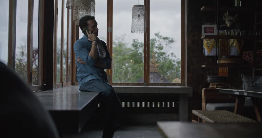 A man is sitting on a counter near windows talking on his cell phone