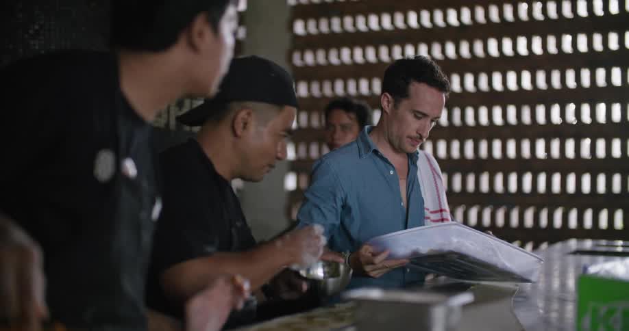 An employee in a restaurant prepares food as a boss looks in a pan and instructs other employees