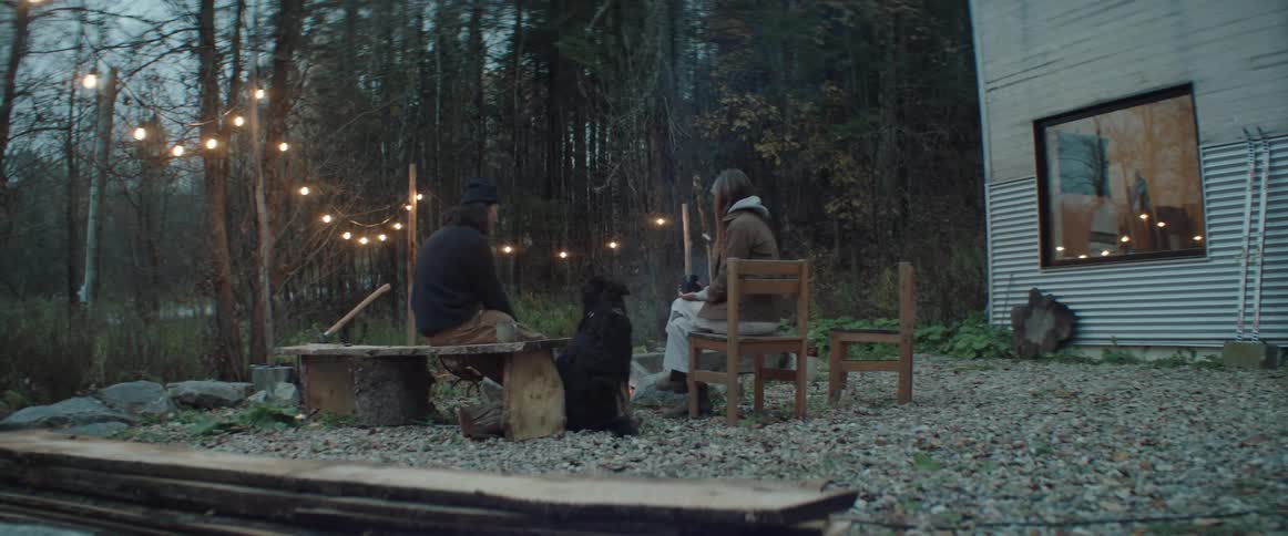 A man pets his dog as he sits outside a home in a rural area with a woman in front of a firepit
