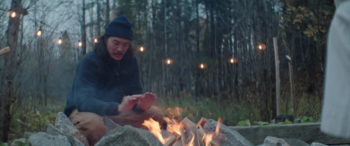 A man crouching next to a campfire and warming his hands